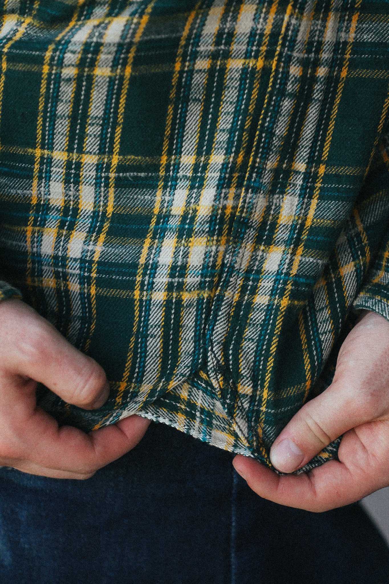 A close-up of a person wearing a green, yellow, and white plaid flannel shirt, holding the fabric at the waist to show its texture and weight.