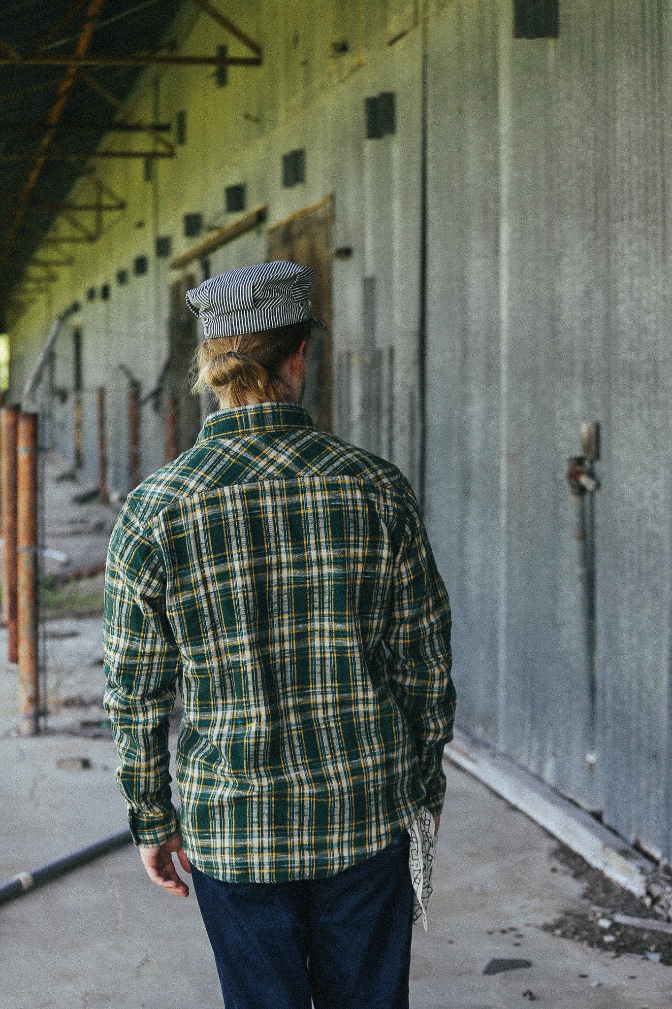 A man viewed from behind wearing a green and yellow plaid flannel shirt, dark blue pants, and a gray flat cap, standing in a concrete corridor next to a large gray metal building.