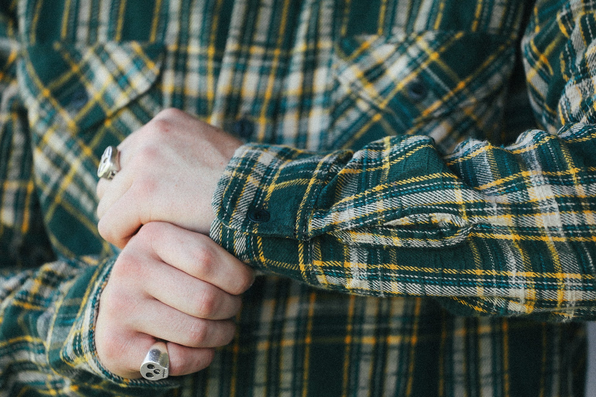 A close-up of a person's hands clasped in front of a green and yellow plaid flannel shirt. The person is wearing silver rings on both fingers.