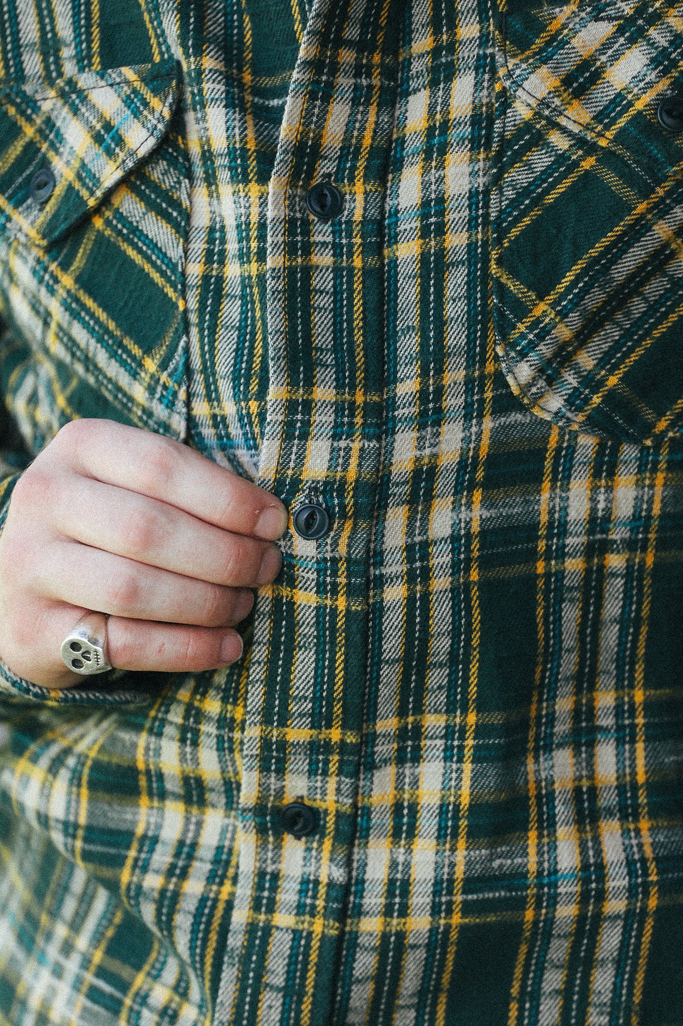 Close-up of a person wearing a green, yellow, and white plaid flannel shirt, with a hand buttoning the front and a skull ring on one finger.