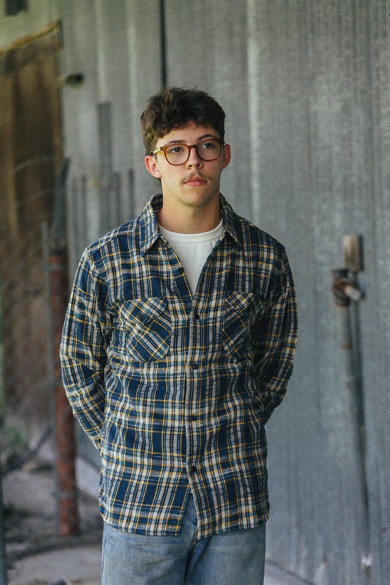 A young man with glasses stands in front of a weathered gray concrete wall, wearing a blue, yellow, and white plaid button-up shirt over a white t-shirt and blue jeans.