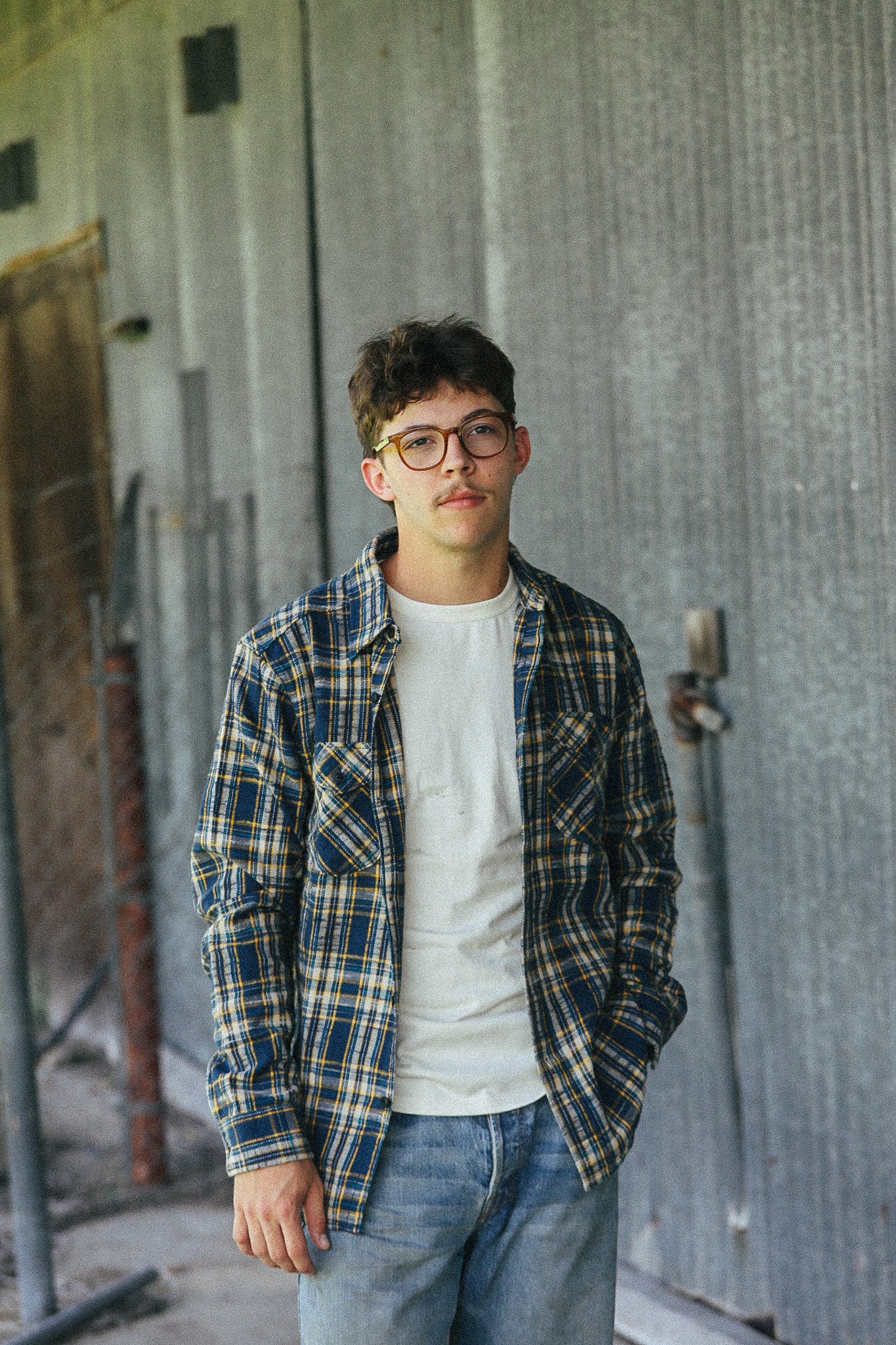A young man wearing round eyeglasses, a blue and yellow plaid flannel shirt over a white t-shirt, and blue jeans, standing in front of a corrugated metal wall.
