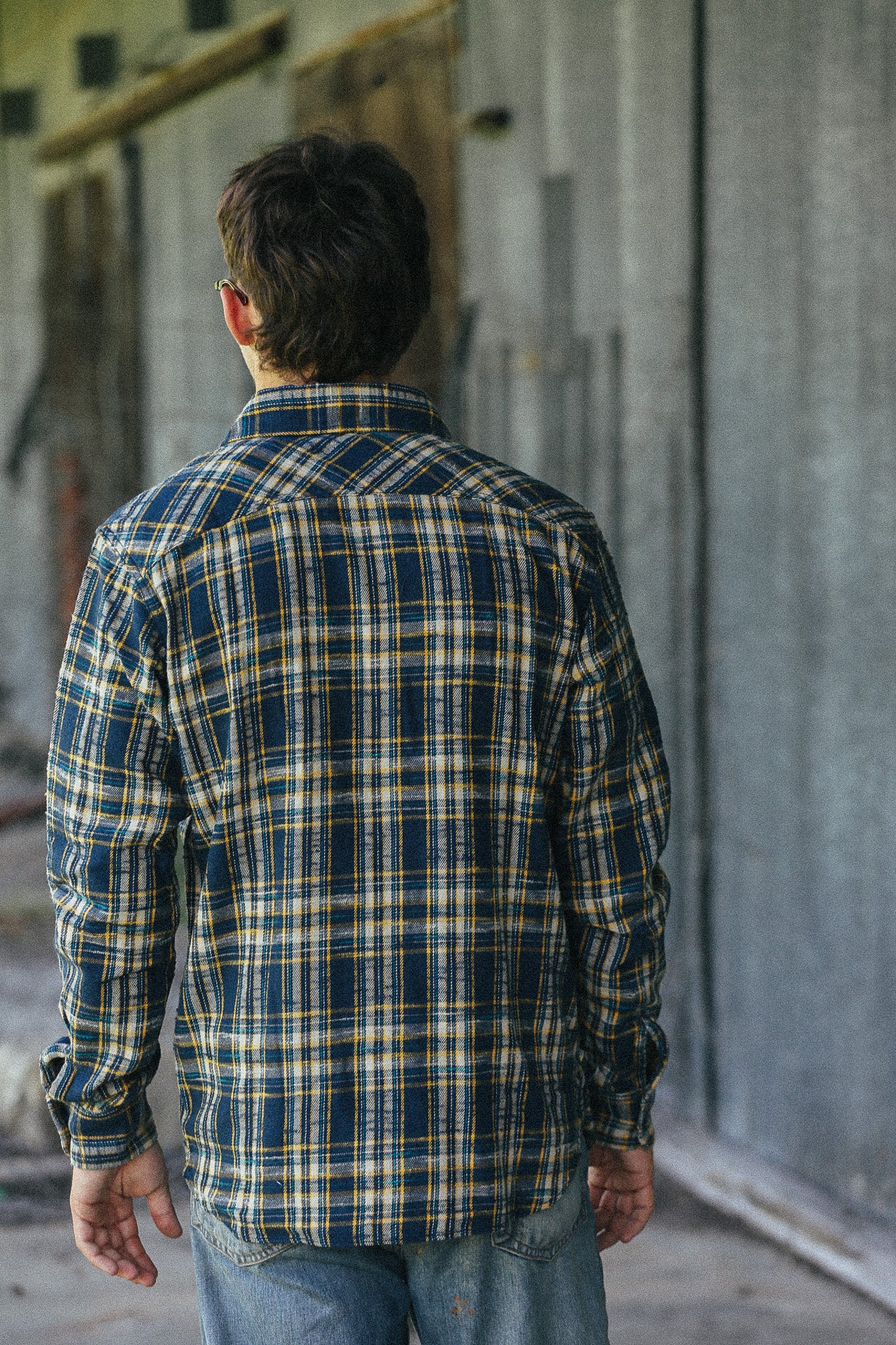 A man viewed from behind wearing a navy blue, yellow, and white plaid flannel shirt and blue jeans, standing in an industrial concrete hallway.