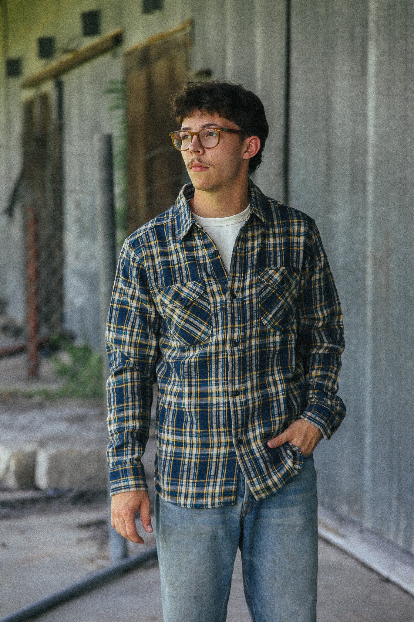 A man wearing round eyeglasses, a blue and yellow plaid button-down shirt over a white t-shirt, and blue jeans, standing in front of a gray corrugated metal wall.