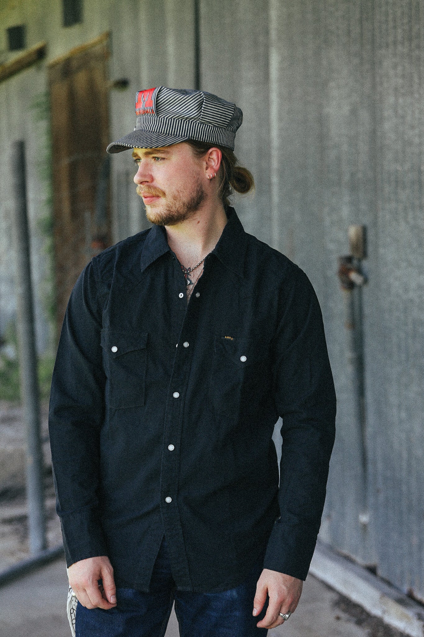 A man wearing a black long-sleeve button-up shirt with silver snap buttons and chest pockets, paired with a gray and black striped cap, standing in front of a gray corrugated metal wall.