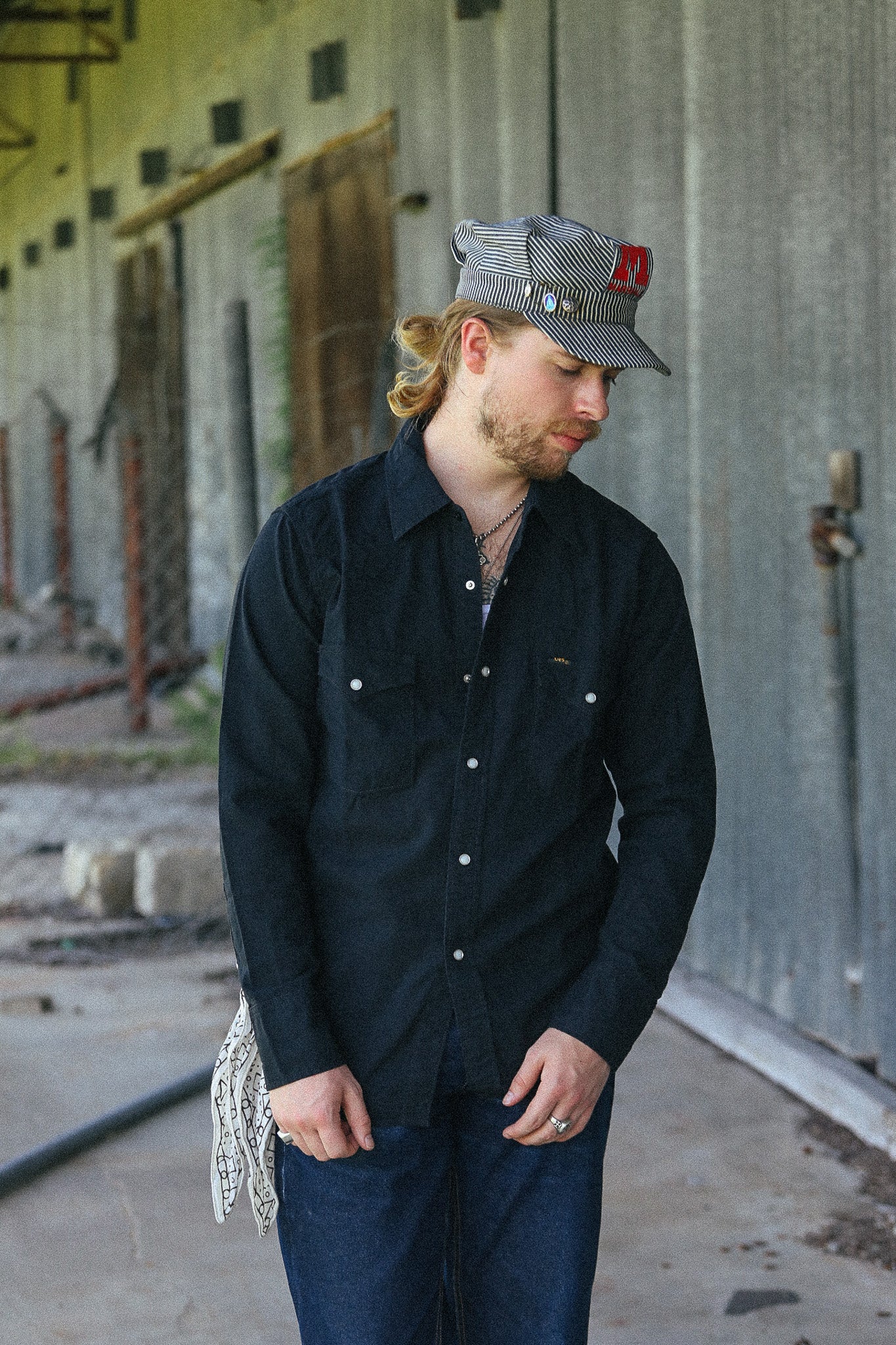 A man wearing a black long-sleeve button-up shirt, dark blue jeans, and a gray patterned cap with a red logo, standing in an industrial concrete corridor.