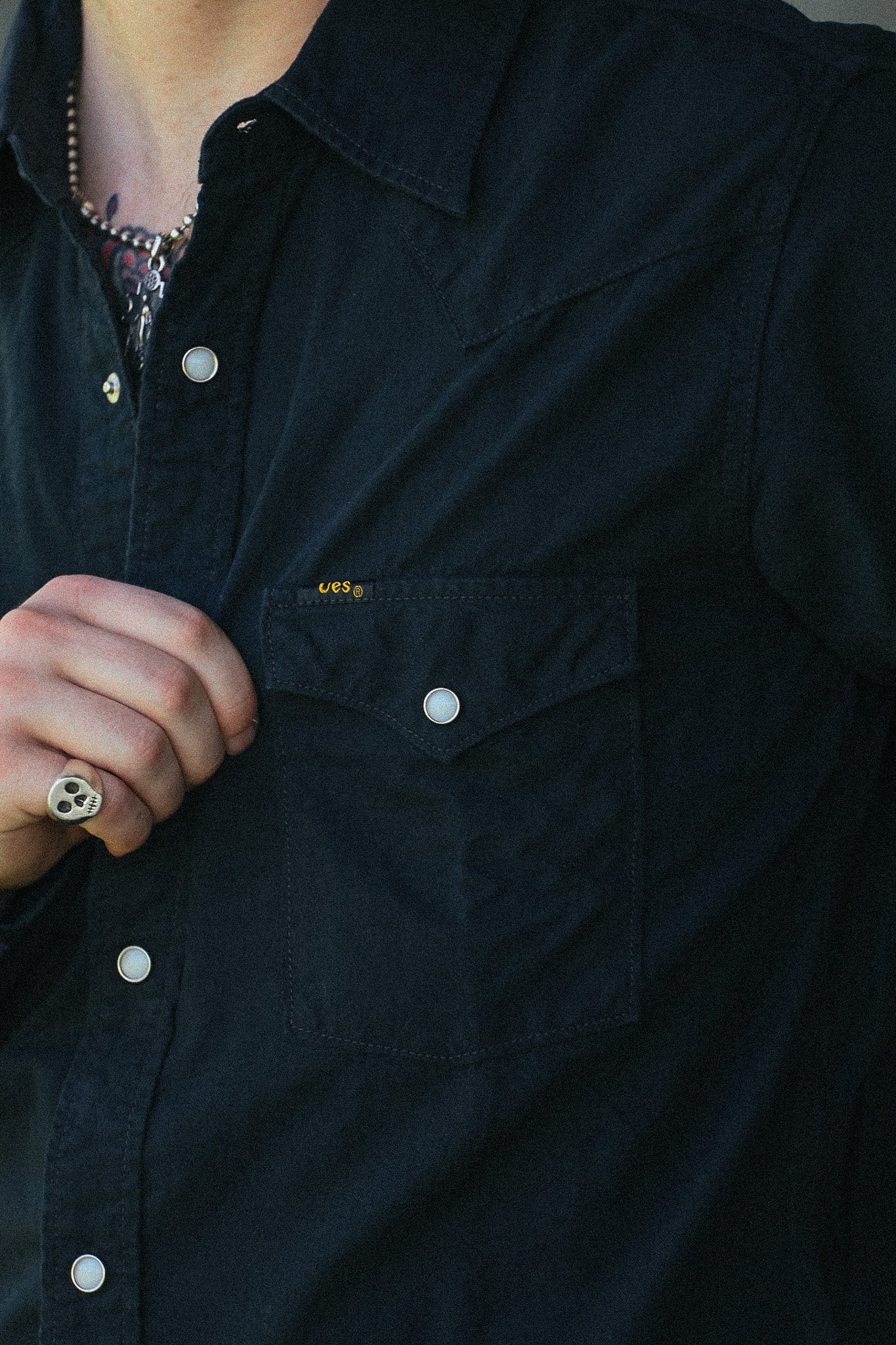 Close-up of a person wearing a dark navy button-up shirt with a chest pocket and white snap buttons. A small yellow embroidered logo is visible on the pocket.