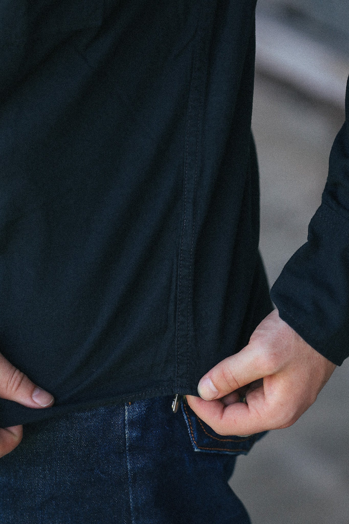 Close-up of a person wearing a black long-sleeve top and dark blue jeans, pulling the hem of the shirt slightly to show the fabric and a small metal zipper detail.