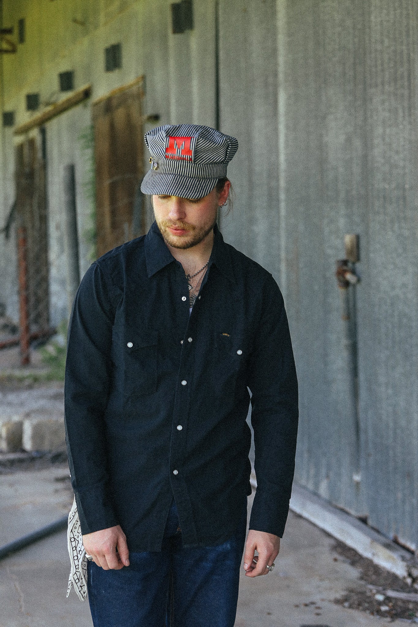 A man wearing a dark navy button-up shirt, blue jeans, and a gray striped cap with a red square patch, standing in front of a weathered concrete wall.