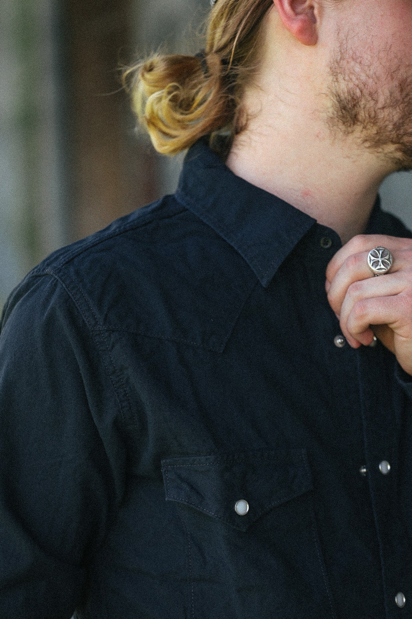 Close-up of a man wearing a dark navy blue denim shirt jacket, featuring a button-down collar, chest pocket with a silver snap, and a ring on his finger.