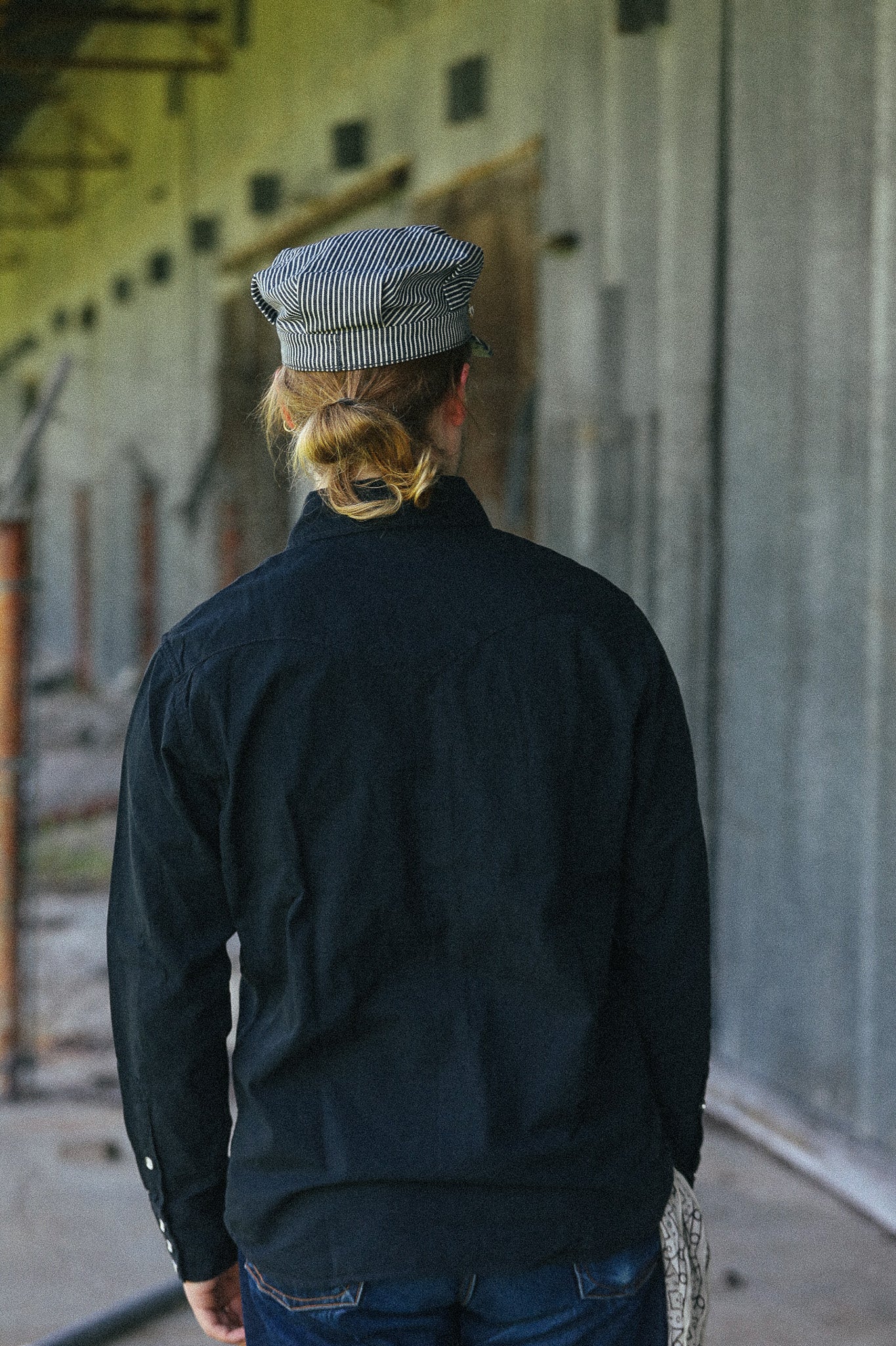 A person viewed from behind wearing a dark navy long-sleeve shirt and a gray-and-white striped cap, standing in a corridor with a concrete wall.