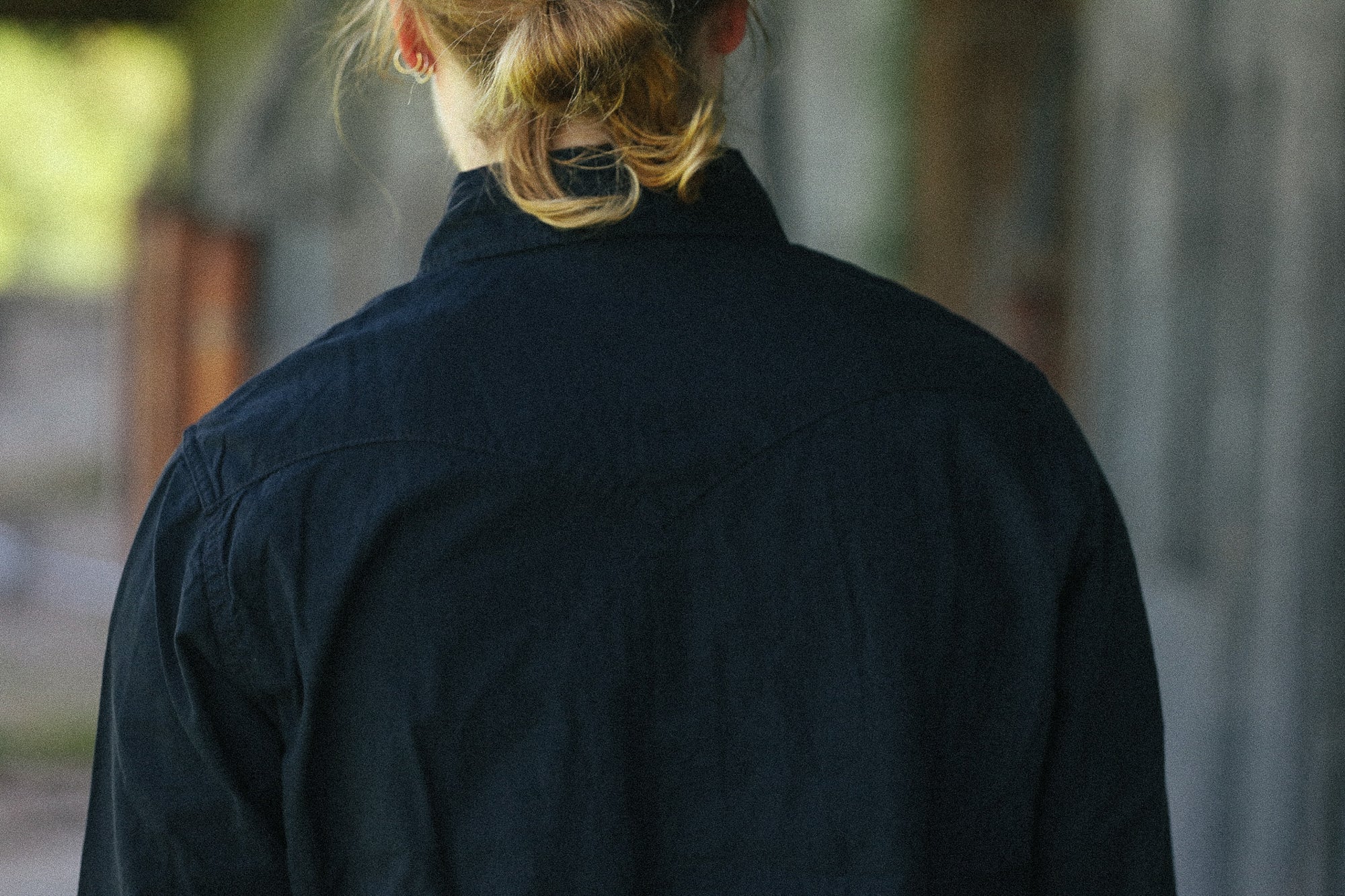 A person viewed from behind wearing a dark navy blue collared shirt or jacket in an outdoor setting with a blurred background.