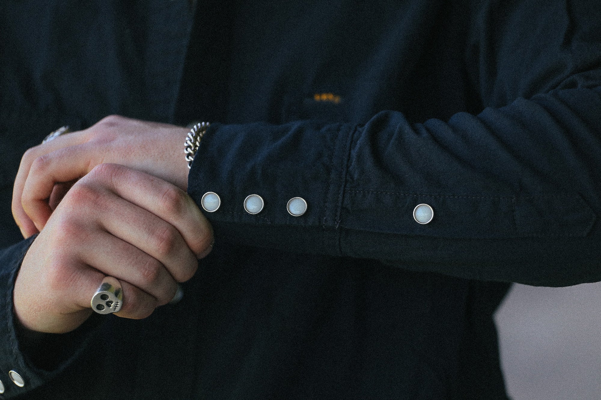 Close-up of a person's hands adjusting a silver chain bracelet on their wrist, wearing a black long-sleeve button-up shirt with white snap buttons on the cuff.