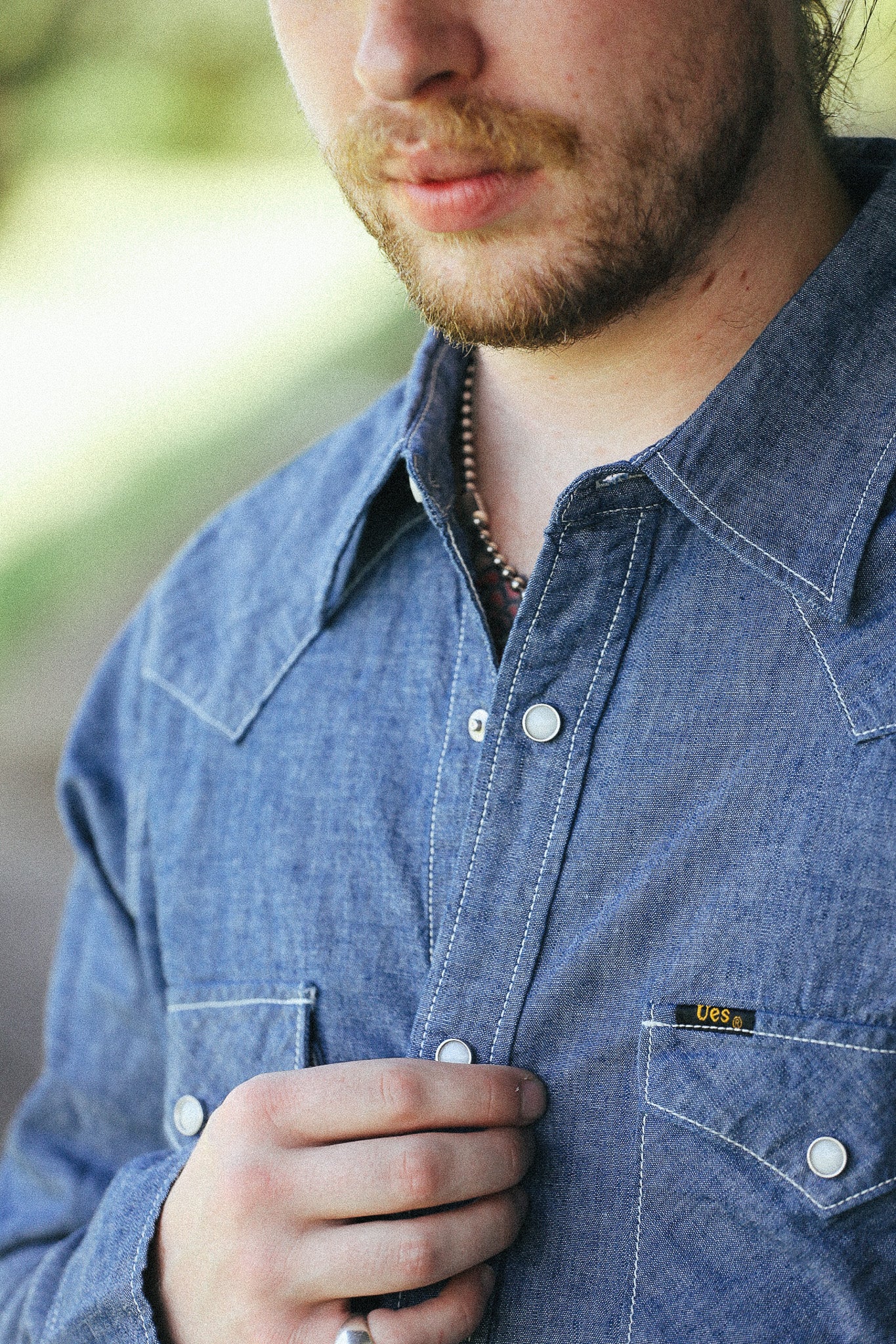 Close-up of a man wearing a blue denim button-up shirt with white contrast stitching and a small yellow Levi's logo patch on the chest pocket.