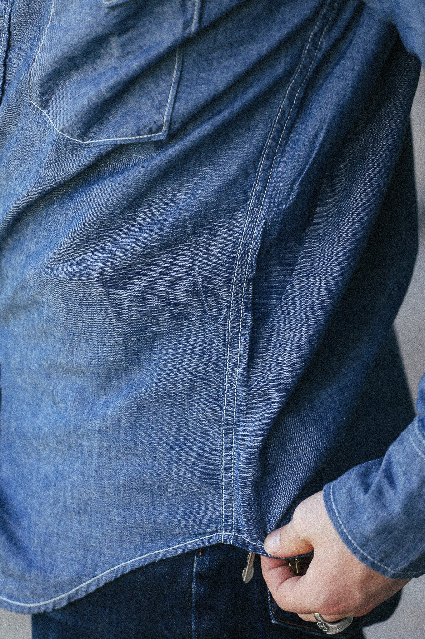 Close-up of a person wearing a blue denim shirt, showing the fabric texture, pocket, and hand adjusting the hem.