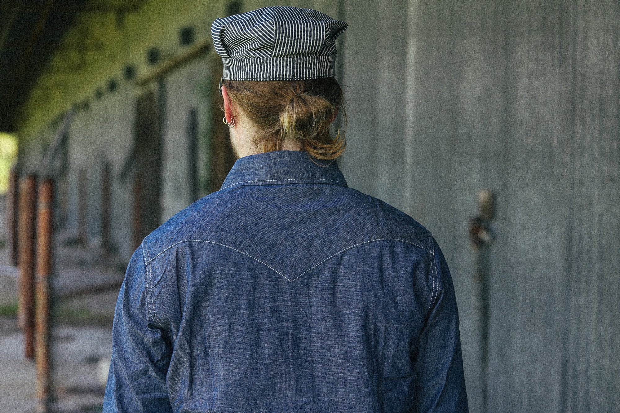 A person viewed from behind wearing a blue denim work shirt and a gray striped cap, standing in a rustic outdoor setting next to a weathered wooden wall.