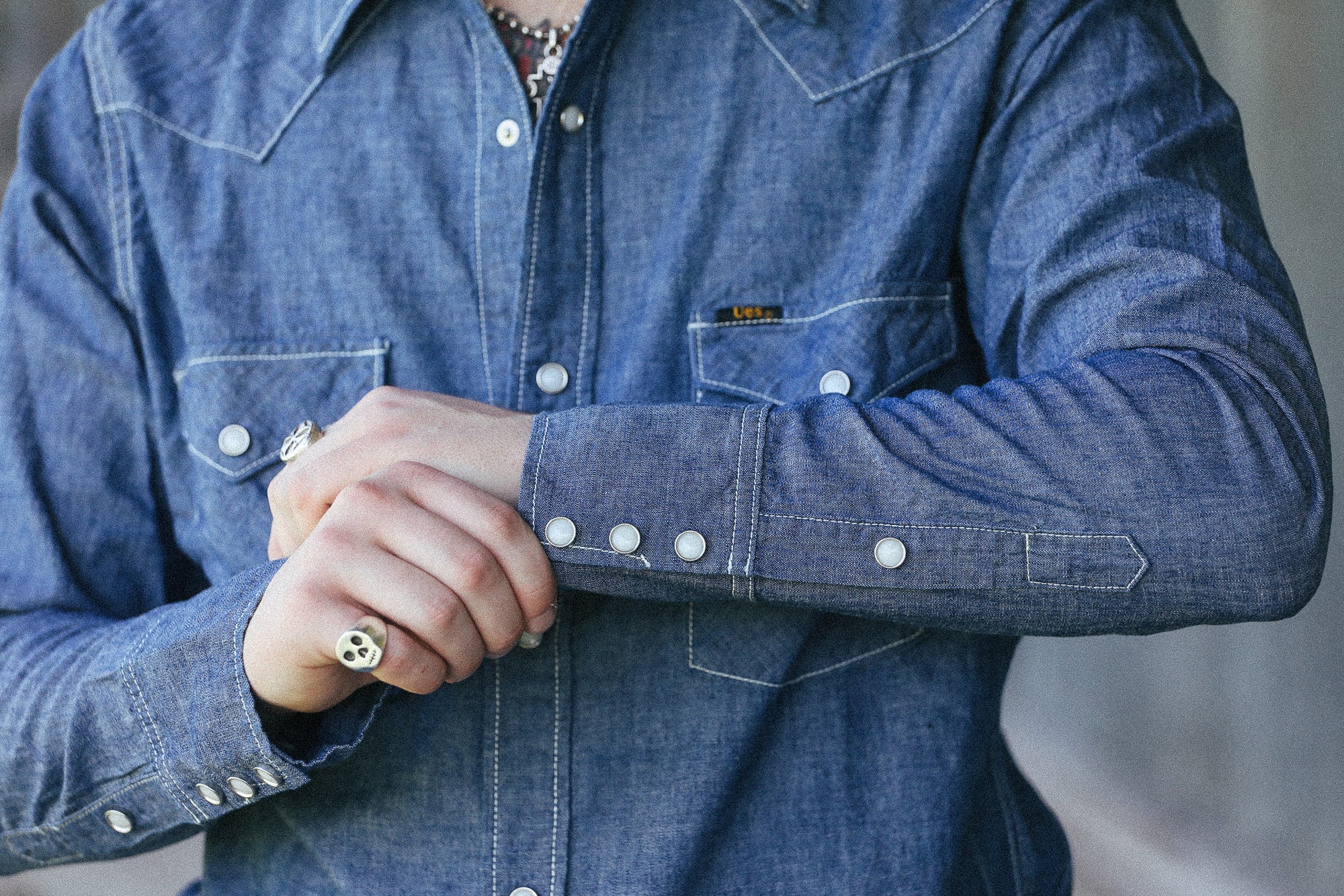A close-up of a person wearing a blue denim long-sleeve western-style button-up shirt, with both hands crossed over the front. The shirt has contrast stitching, silver-toned buttons, and a small chest pocket.