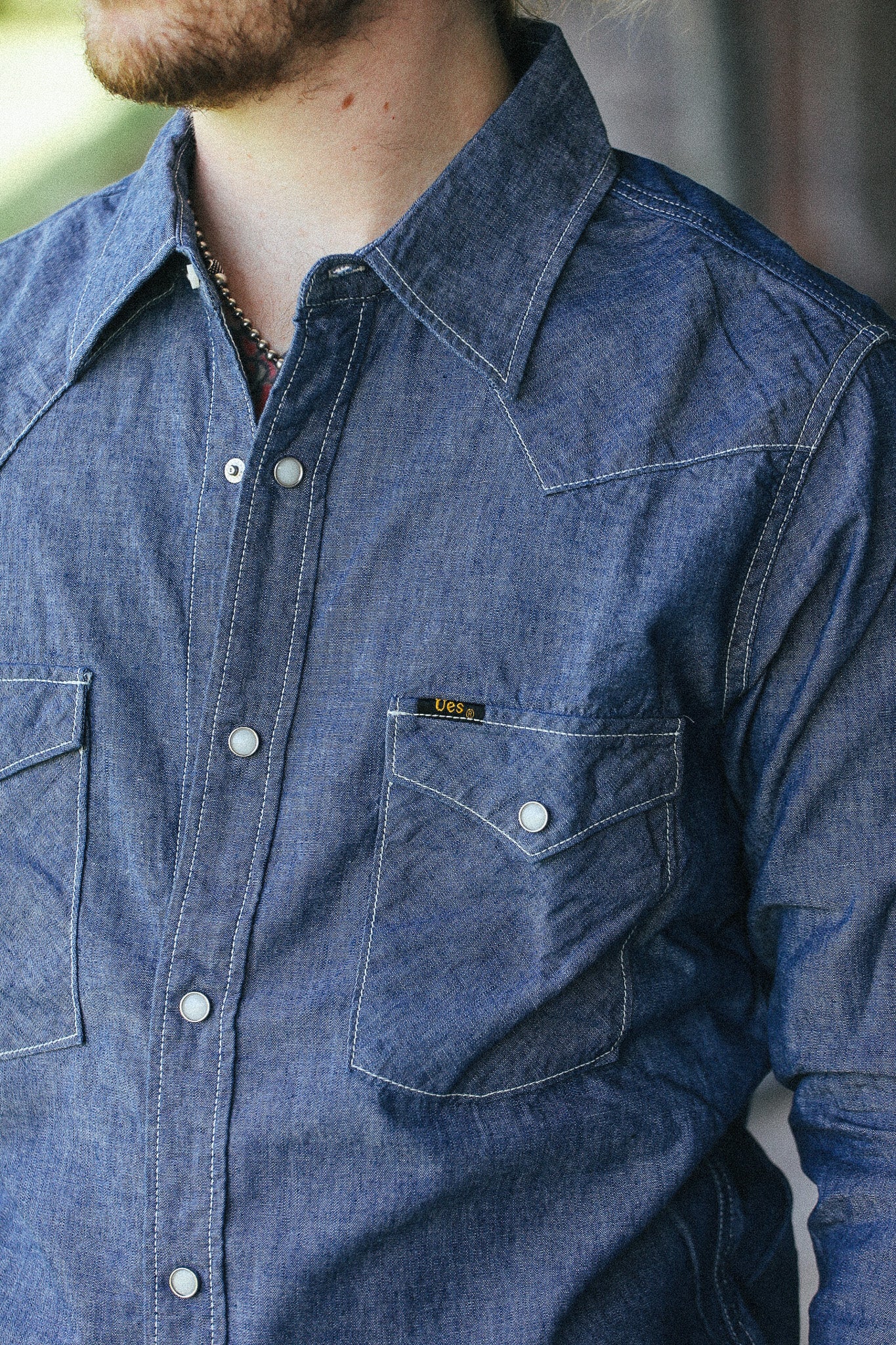 A close-up of a man wearing a blue denim western-style button-up shirt with a chest pocket and a small yellow brand tab.