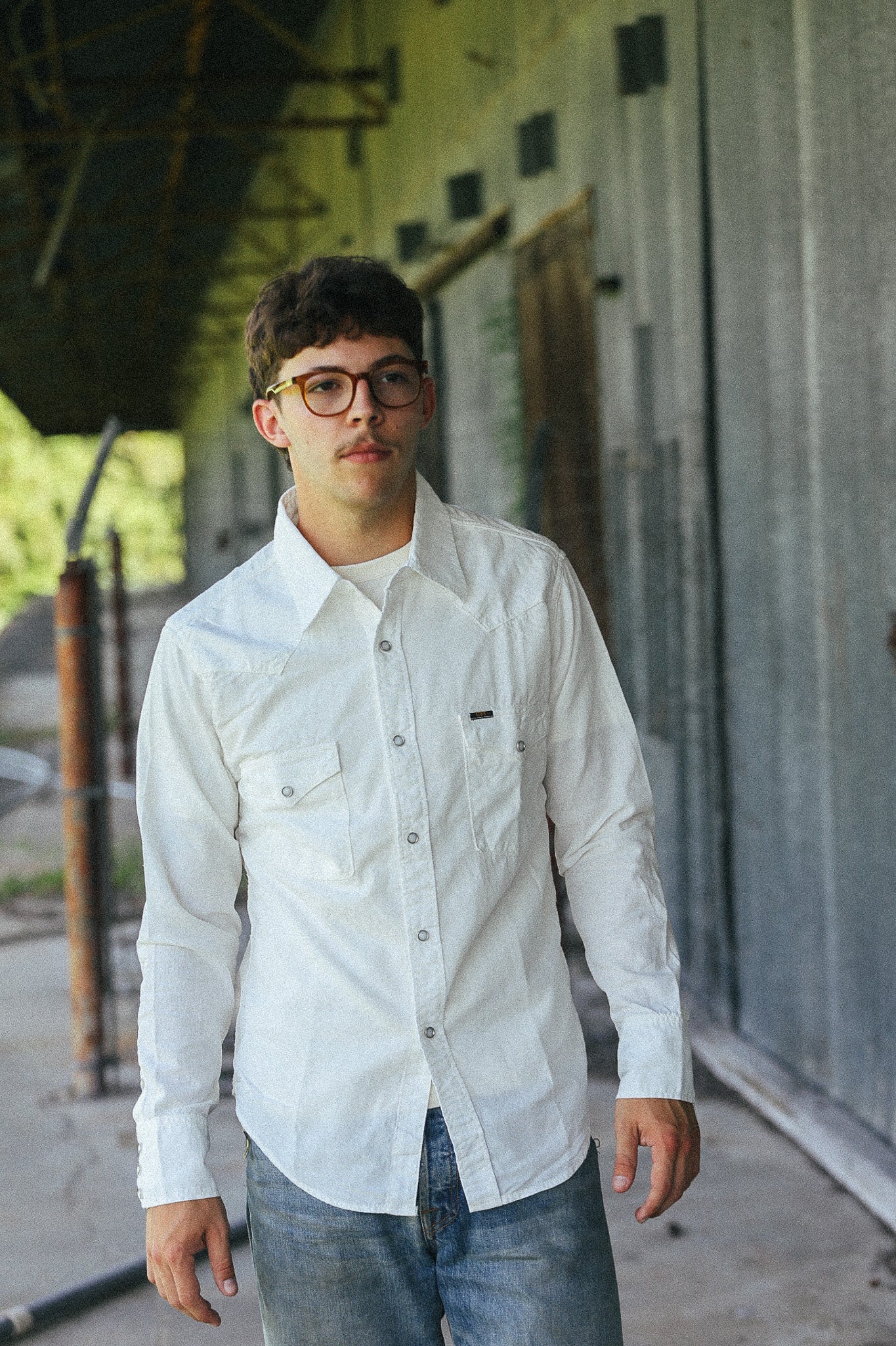 A man wearing a white long-sleeve button-down shirt and blue jeans, standing in front of a rustic gray metal wall.