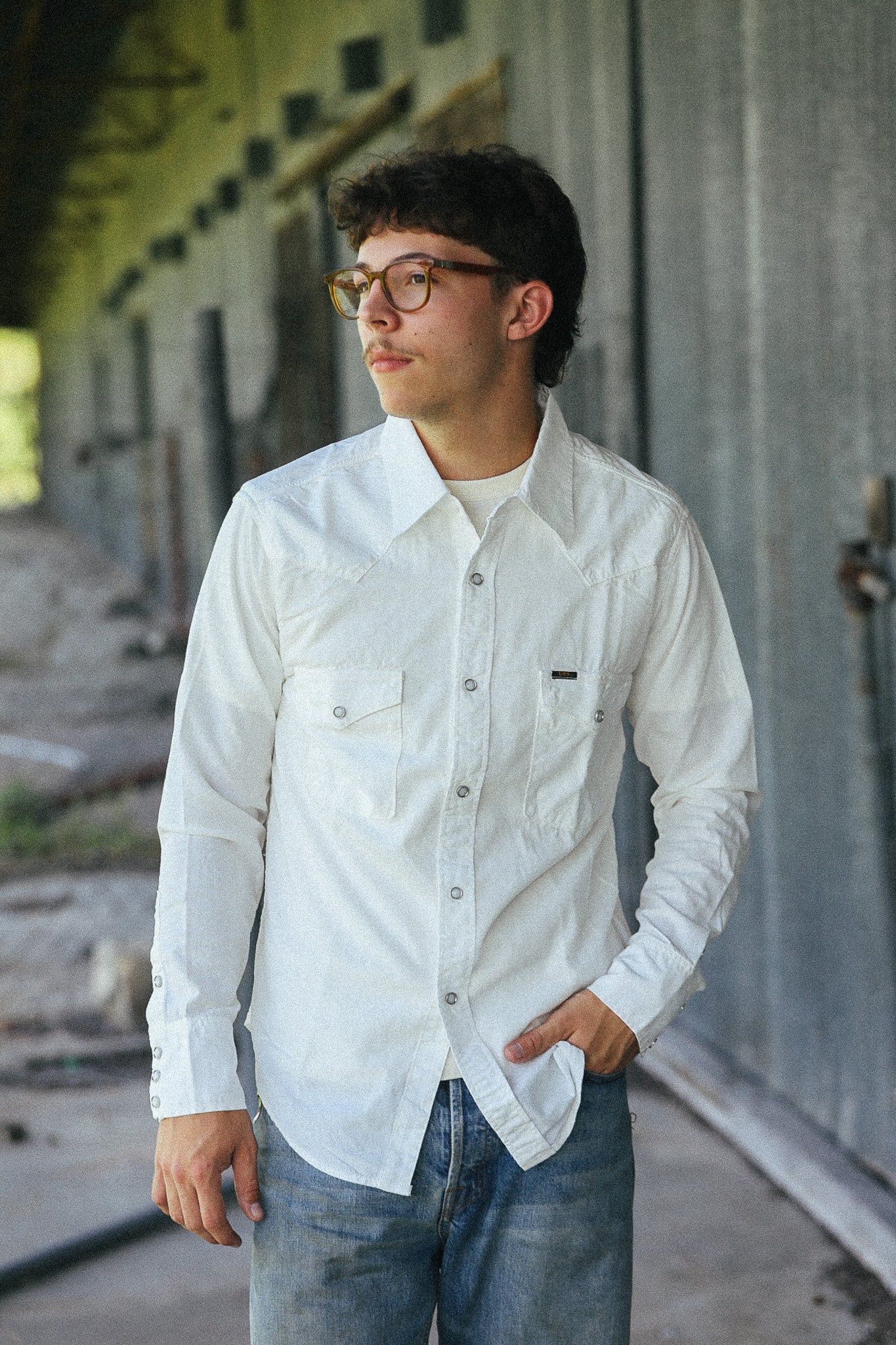 A man wearing a white long-sleeve button-up shirt with two chest pockets and blue jeans, standing in an outdoor industrial setting.