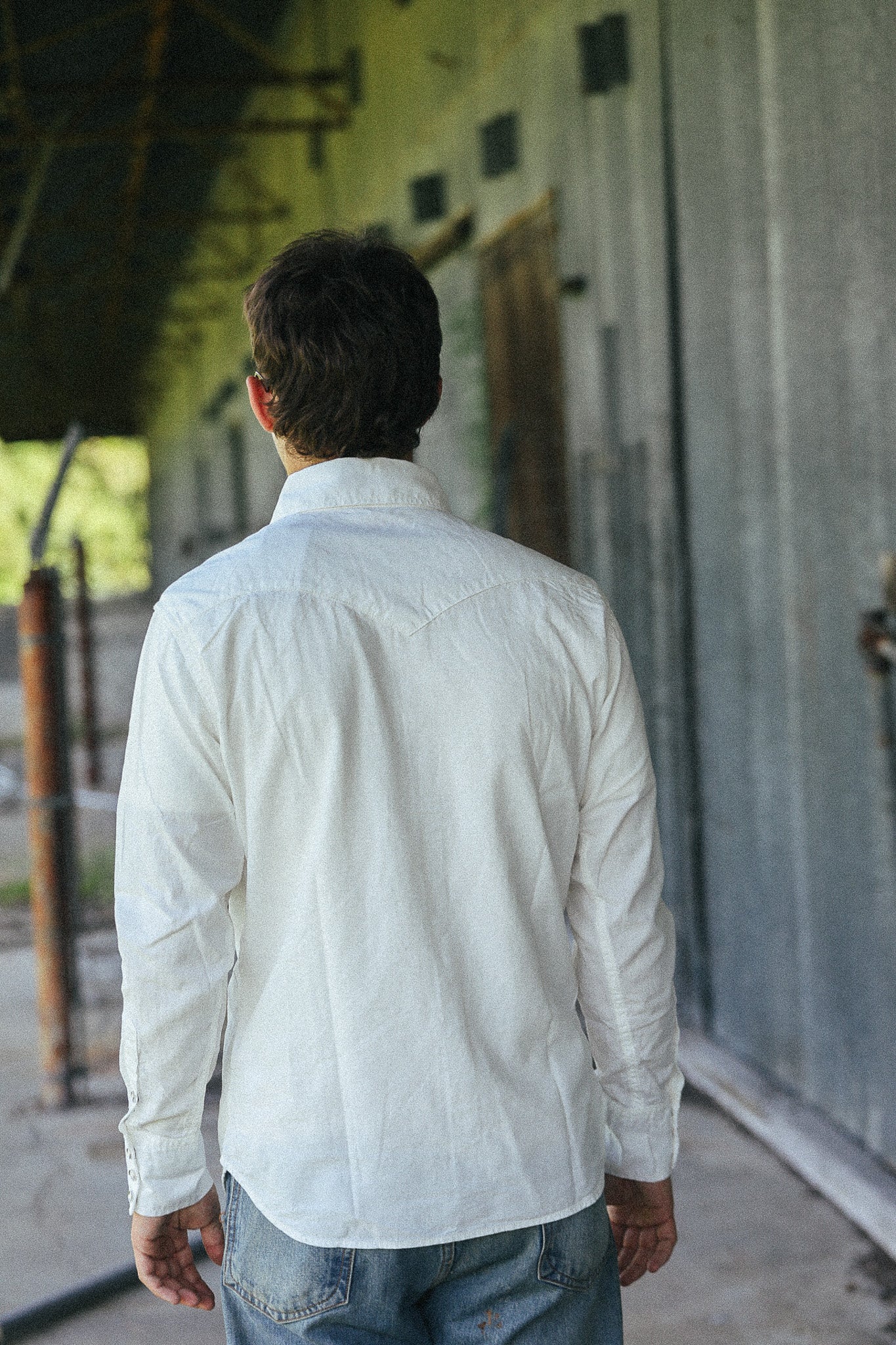 A man viewed from behind wearing a white long-sleeve button-down shirt and blue jeans, standing in a rustic covered walkway next to a corrugated metal wall.
