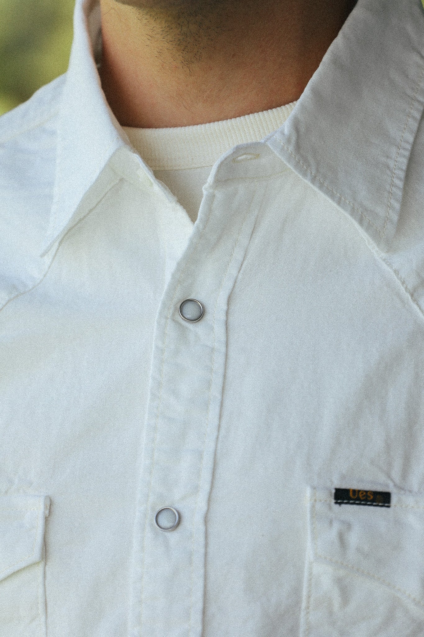Close-up of a person wearing a white button-up shirt with a pointed collar, silver-toned snap buttons, and a small black chest pocket label.