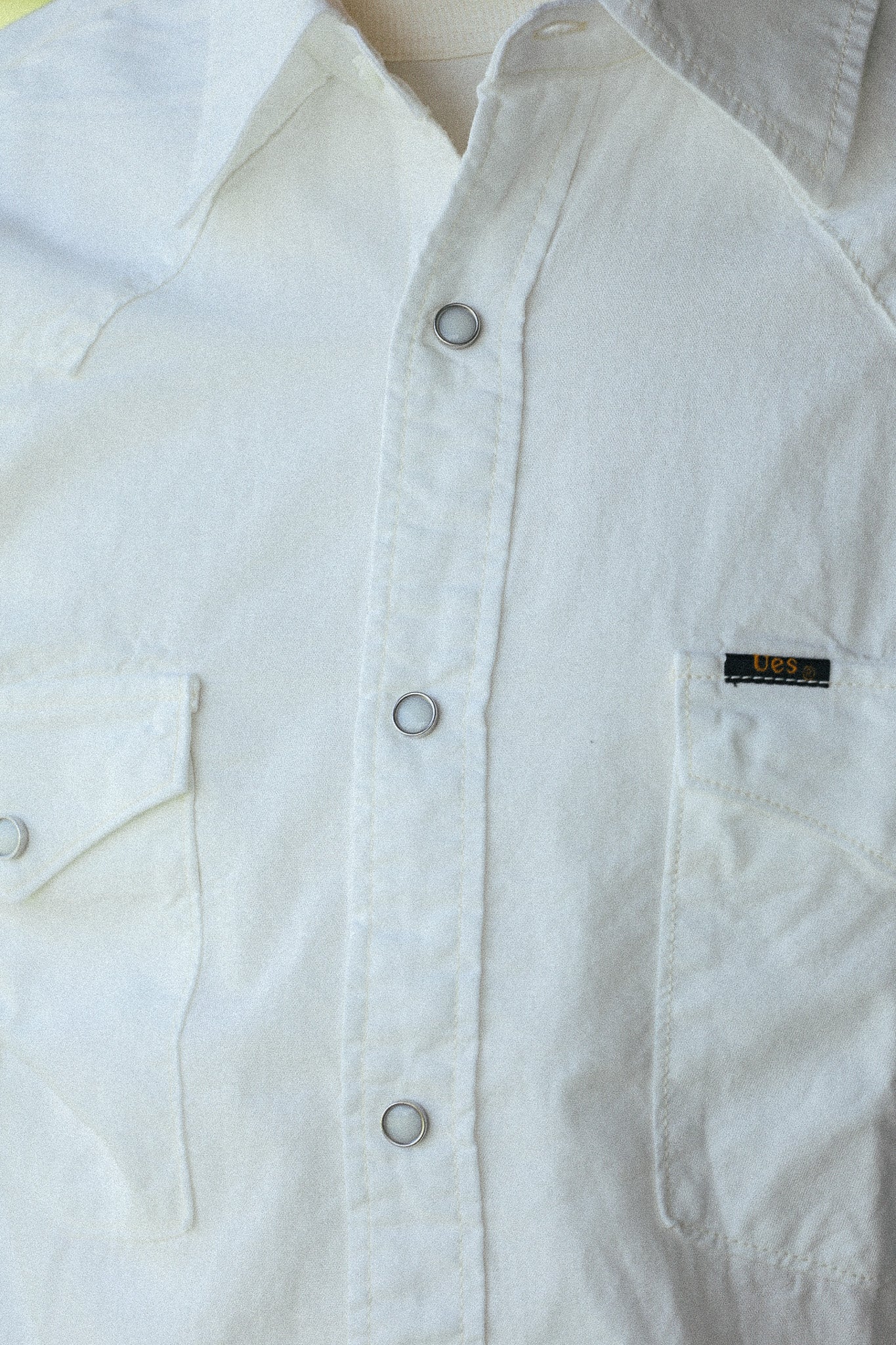 Close-up of a white button-up shirt with silver buttons, a chest pocket, and a small black brand tag on the pocket.