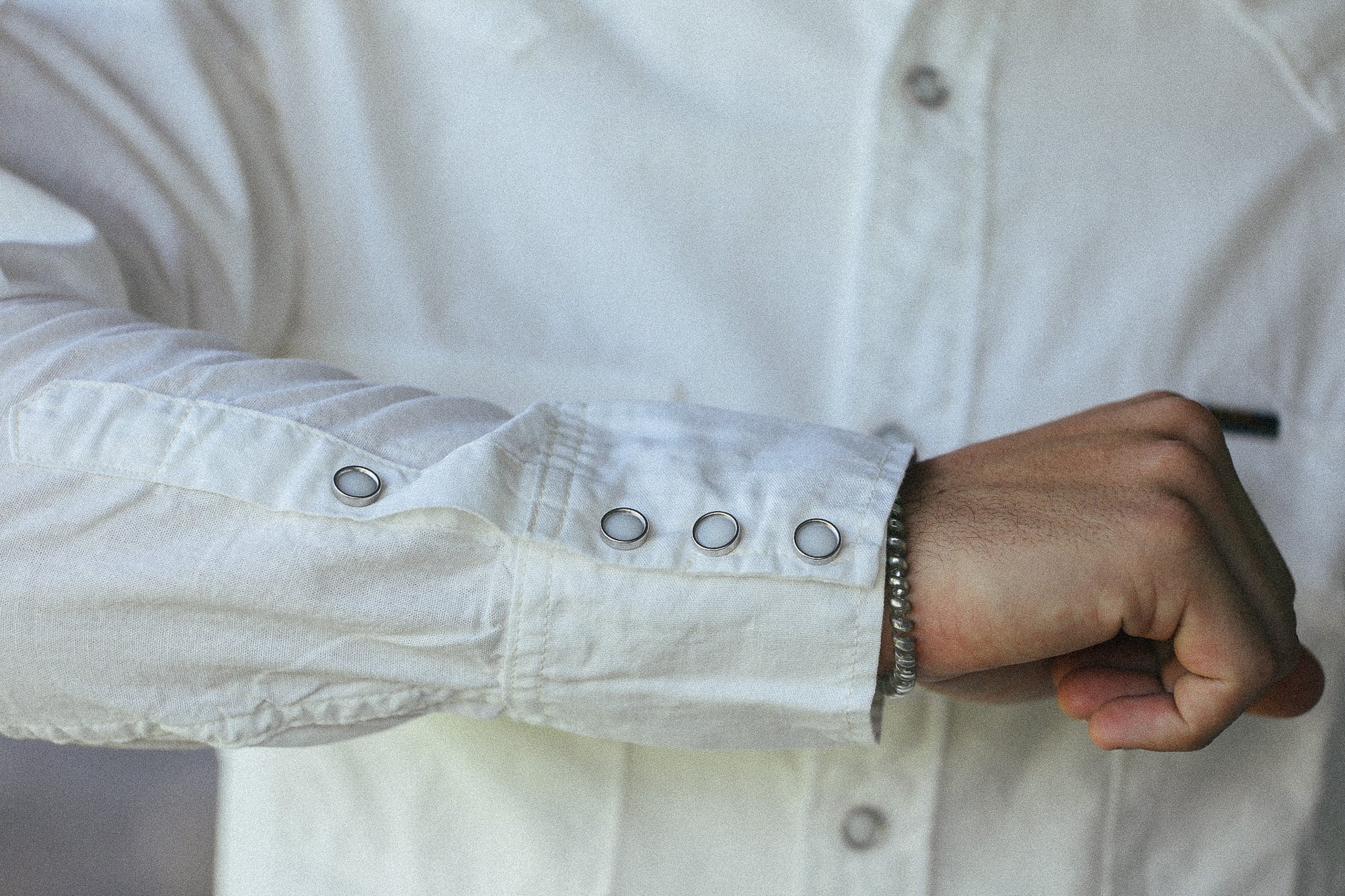 Close-up of a person wearing a white long-sleeve button-up shirt, focusing on the cuff with silver snap buttons and a silver bracelet on the wrist.