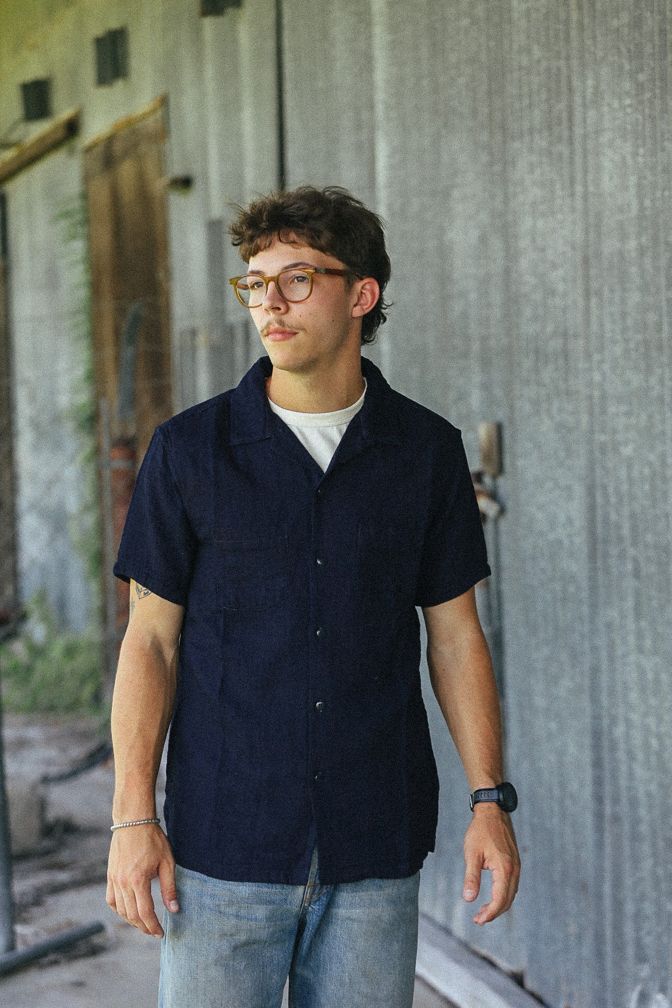 A young man wearing a dark navy blue short-sleeve button-up shirt and light blue jeans, standing in front of a gray corrugated metal wall.