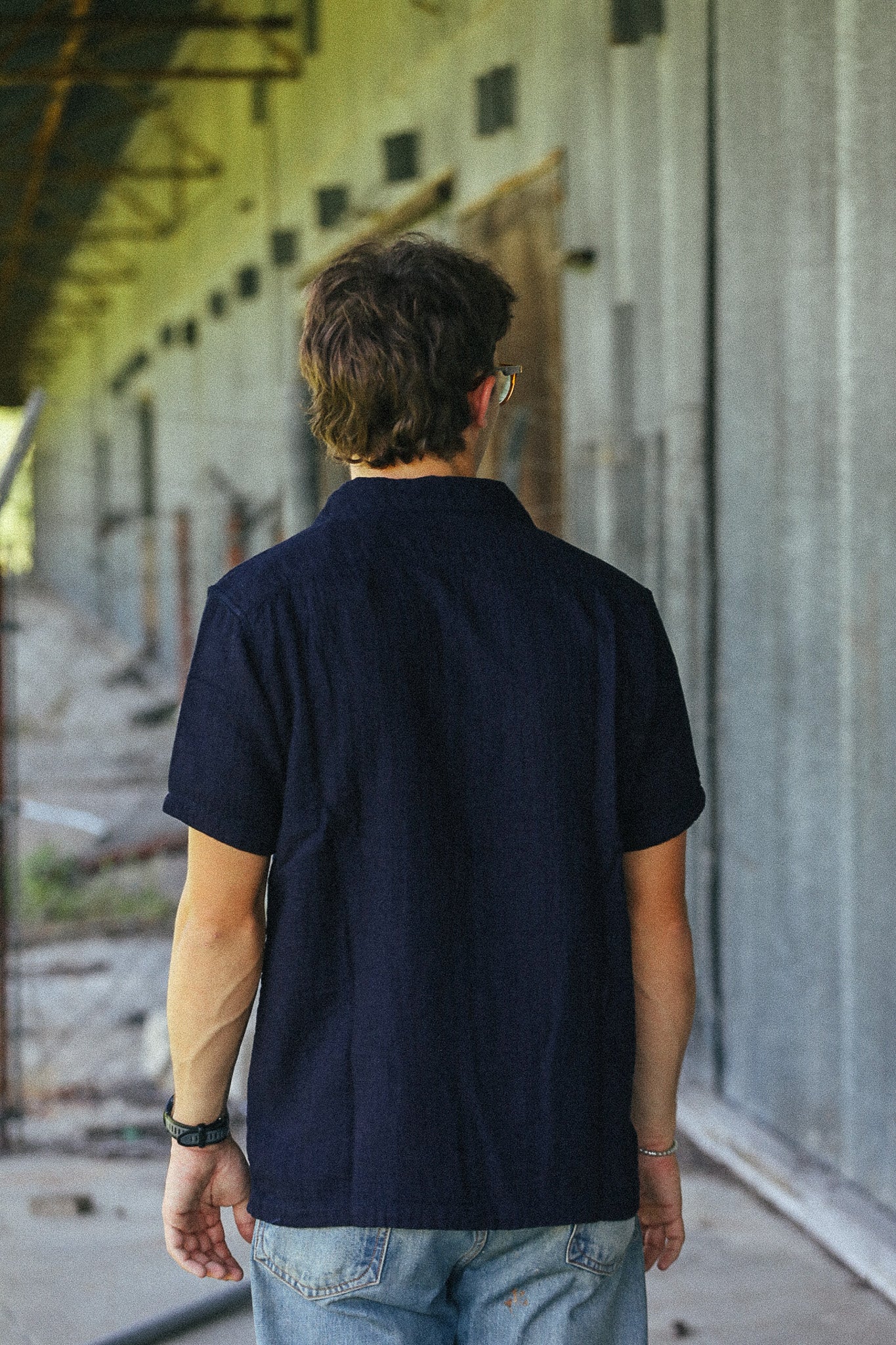 A man viewed from behind wearing a dark navy blue short-sleeve button-up shirt and light blue jeans, standing in an industrial corridor with concrete walls.