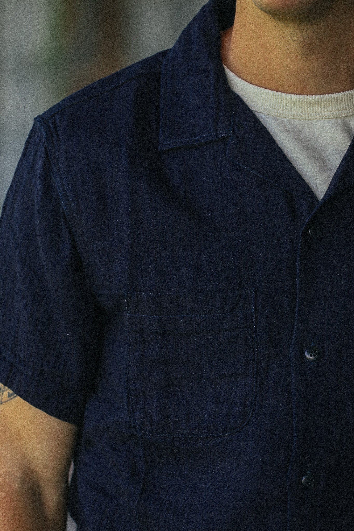 A close-up of a person wearing a dark navy blue short-sleeve button-up shirt with a chest pocket, layered over a white t-shirt.