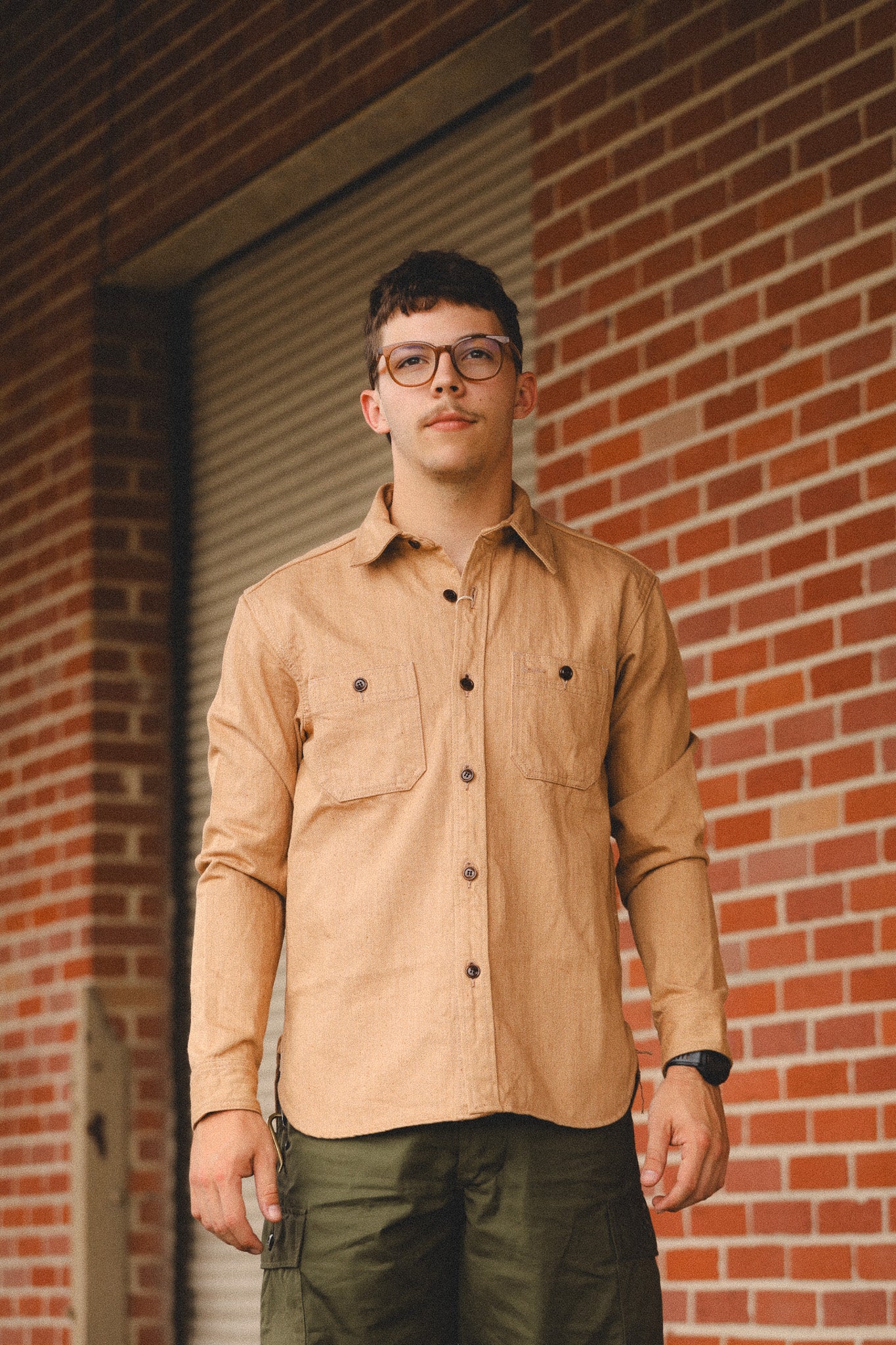 Man wearing a beige shirt and glasses standing in front of a brick wall