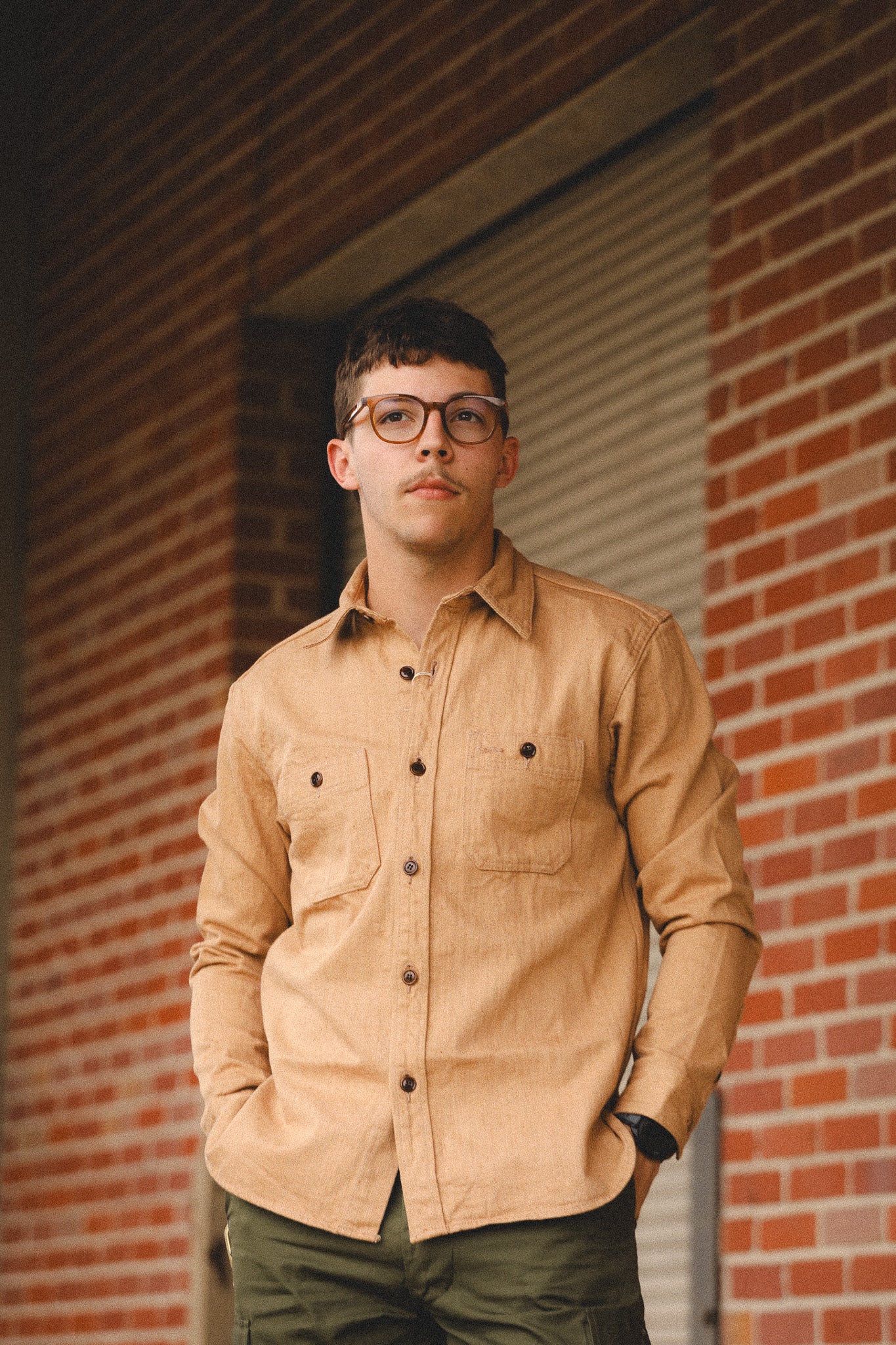 Man wearing a beige shirt and glasses standing against a brick wall.