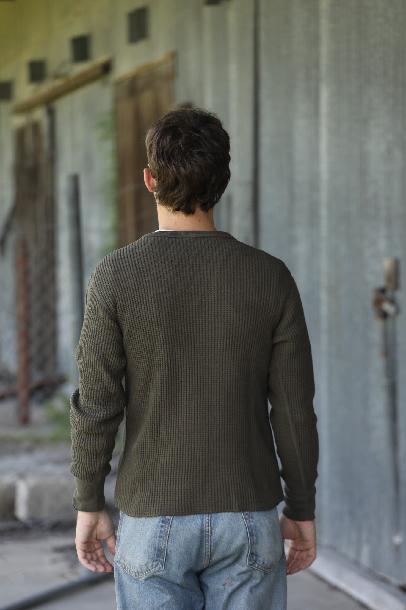 A man viewed from behind wearing an olive green ribbed long-sleeve shirt and light blue jeans, standing in front of a weathered gray metal wall.