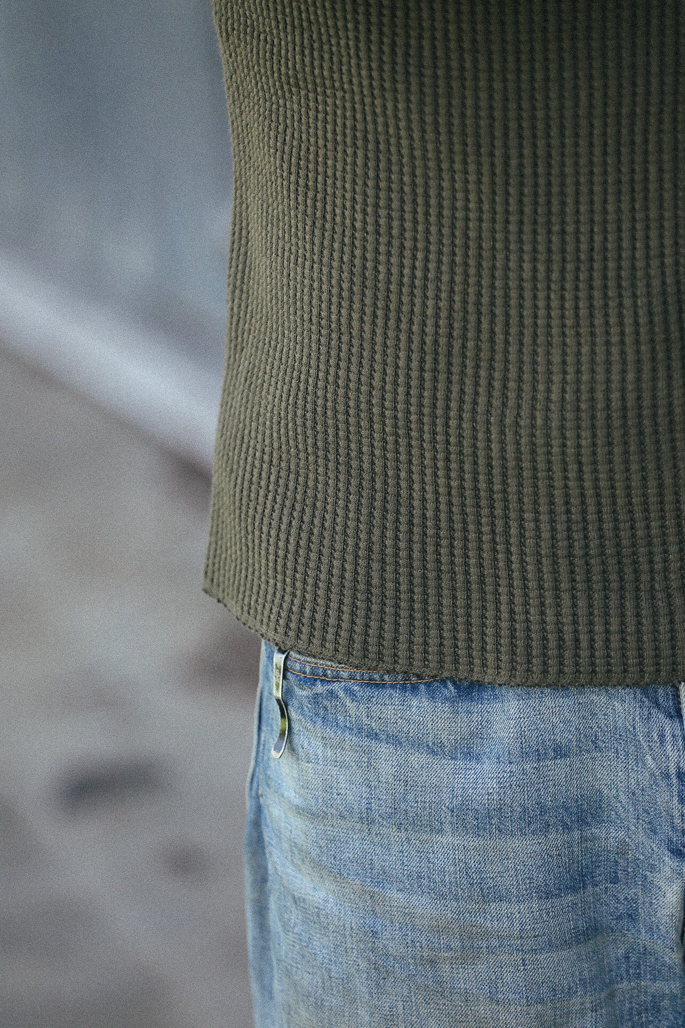 Close-up of a person wearing an olive green ribbed knit top and light blue jeans, cropped from the chest down against a blurred gray background.