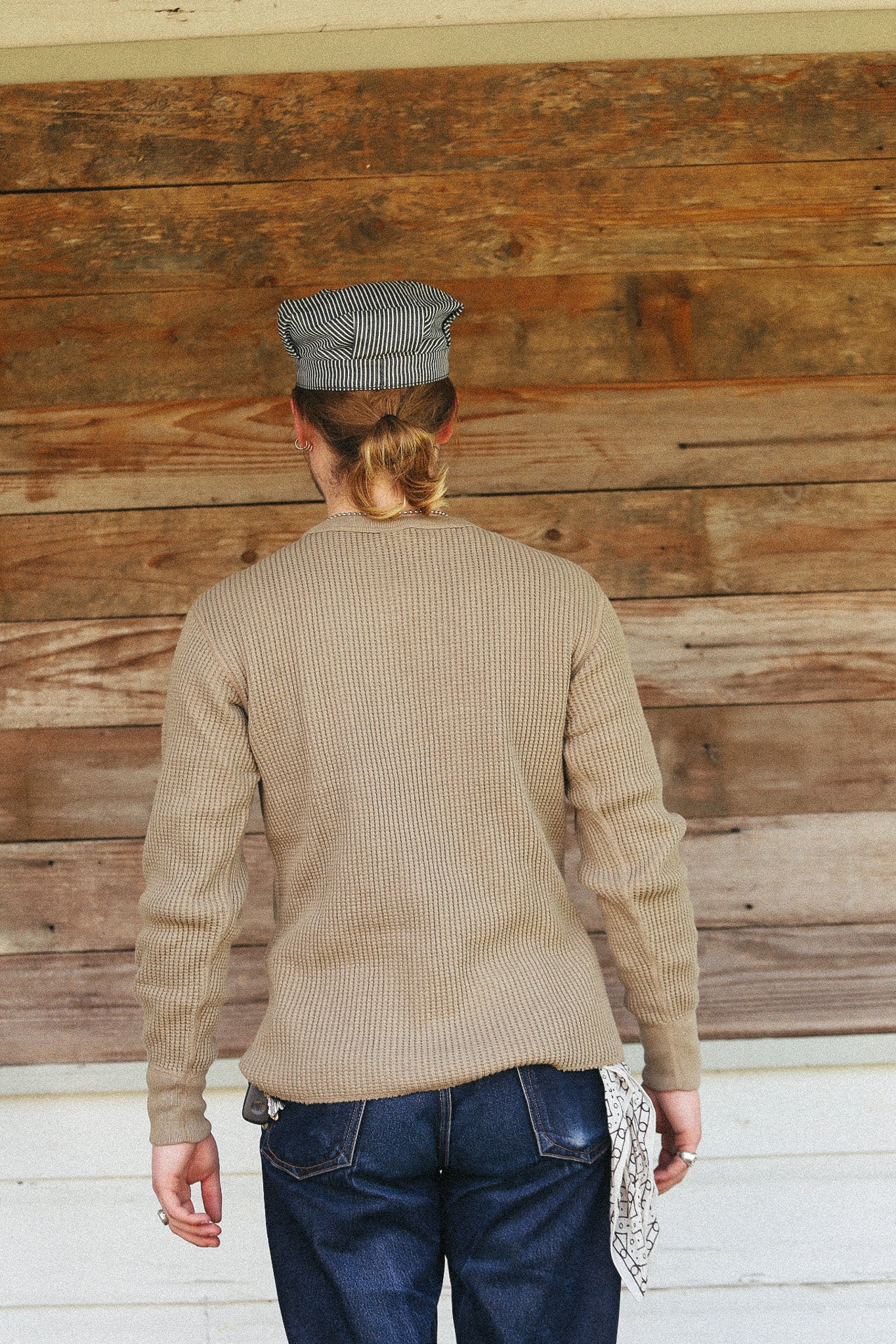 A person viewed from behind wearing a beige ribbed knit sweater, dark blue jeans, and a gray-and-white patterned flat cap, standing in front of a rustic wooden wall.