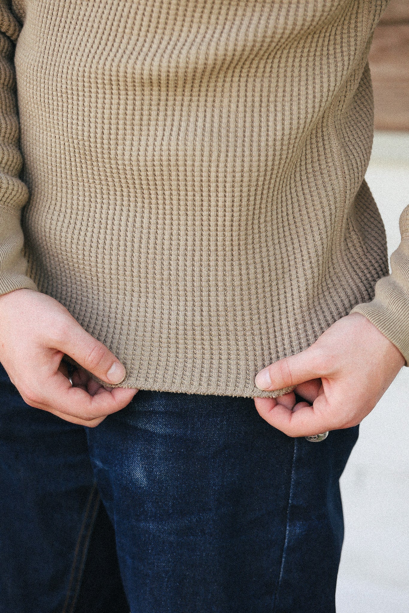 Close-up of a person wearing a beige waffle-knit sweater and dark blue jeans, pulling the hem of the sweater slightly with both hands.