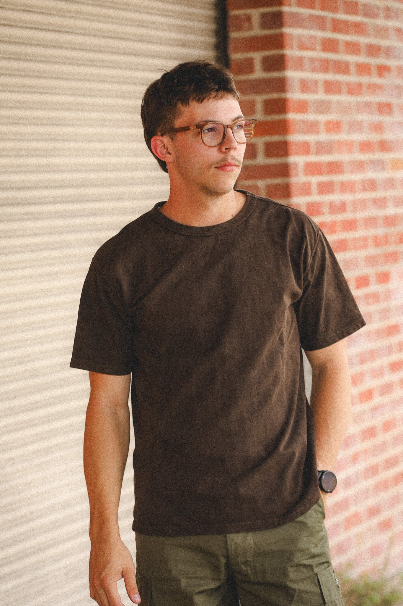 Man wearing a dark brown t-shirt and glasses standing against a brick wall.