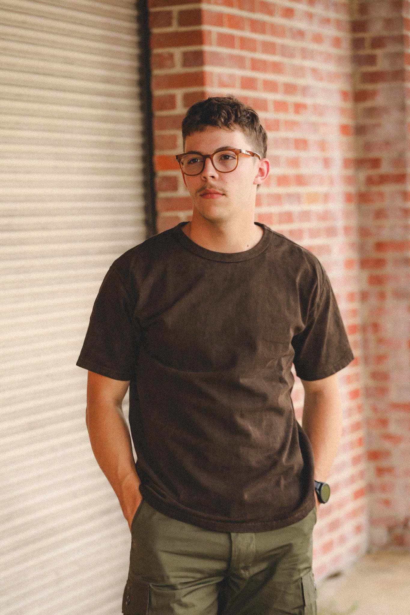 Man wearing a brown t-shirt and glasses standing against a brick wall.