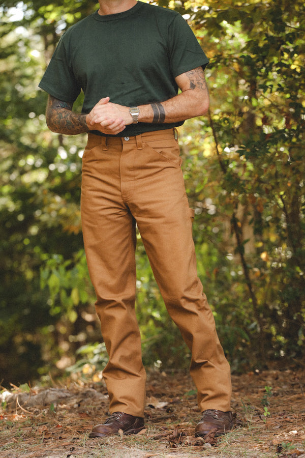 A man wearing brown work pants and a dark green t-shirt stands outdoors in a wooded setting, showcasing the fit of the pants.