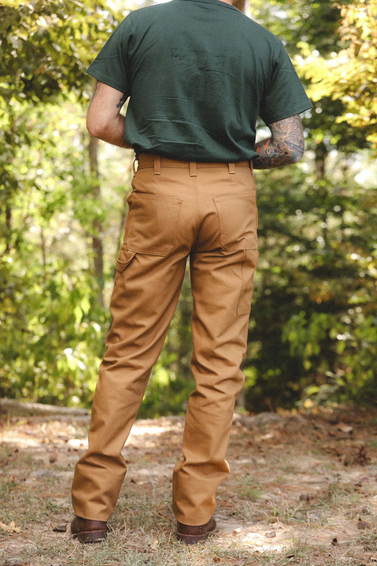 A man viewed from behind wearing tan cargo pants, a dark green t-shirt, and brown boots, standing outdoors in a wooded setting.