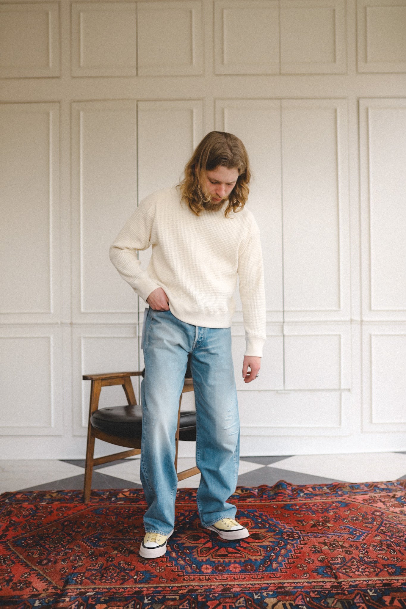 Person wearing a white sweater and blue jeans standing in a room with a chair and patterned rug.