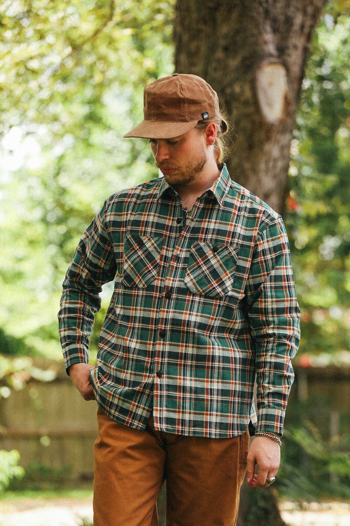 A man wearing a green and brown plaid button-down shirt, brown pants, and a brown cap, standing outdoors in front of a tree.