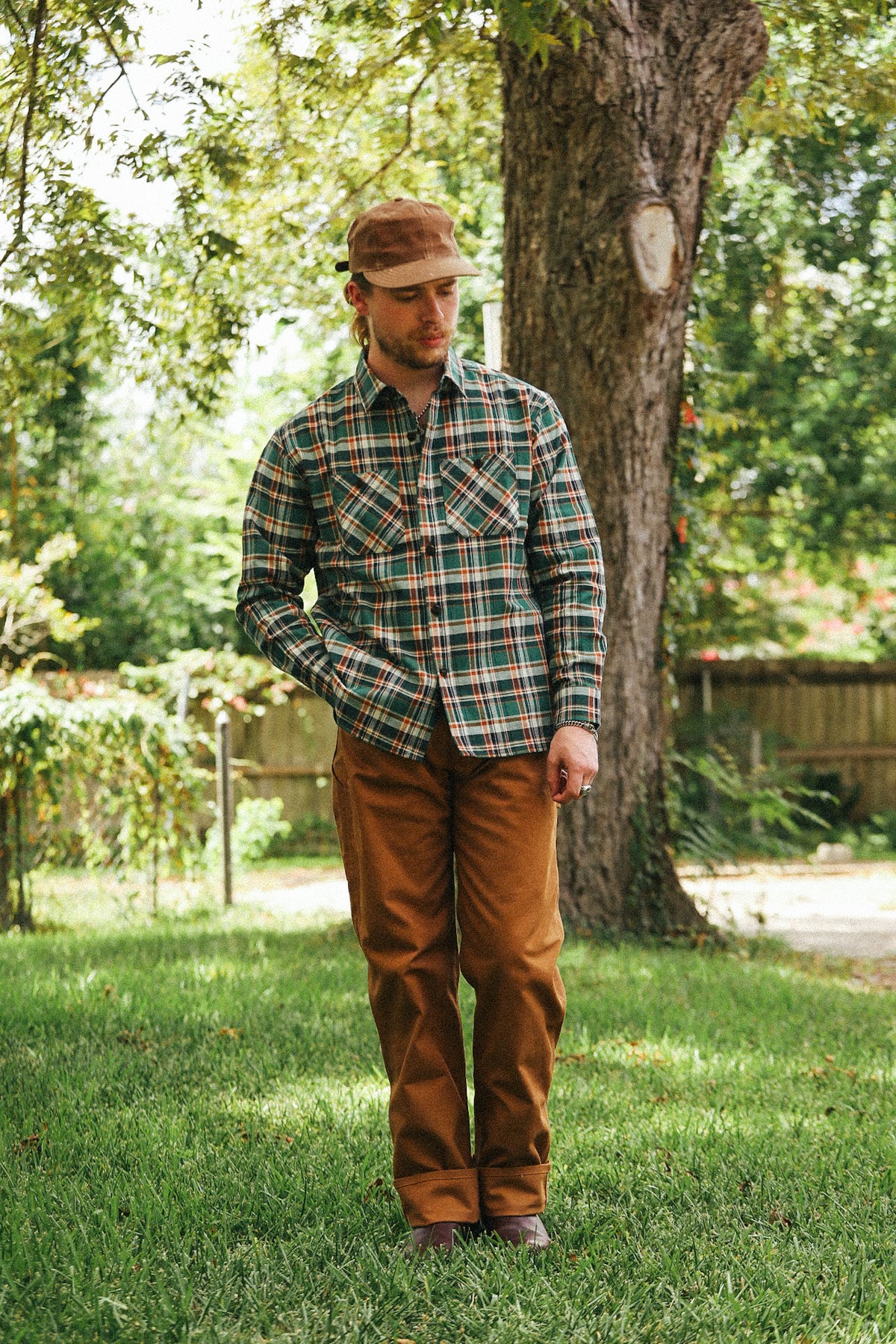A man standing outdoors in a grassy yard next to a large tree, wearing a green and brown plaid long-sleeve button-up shirt, brown pants, and a brown cap.