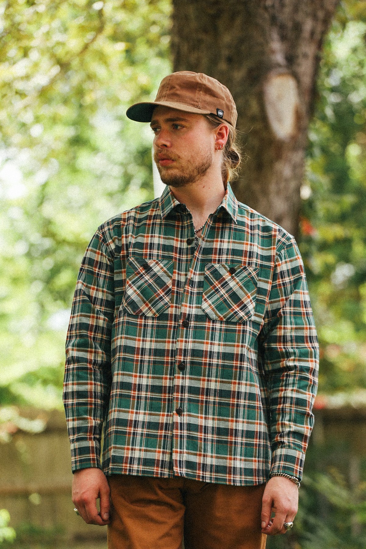 A man wearing a green and brown plaid long-sleeve button-up shirt, brown pants, and a brown cap, standing outdoors in front of a tree.