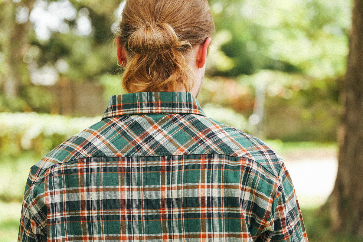 A person viewed from behind wearing a green, orange, and white plaid flannel shirt in an outdoor park setting.