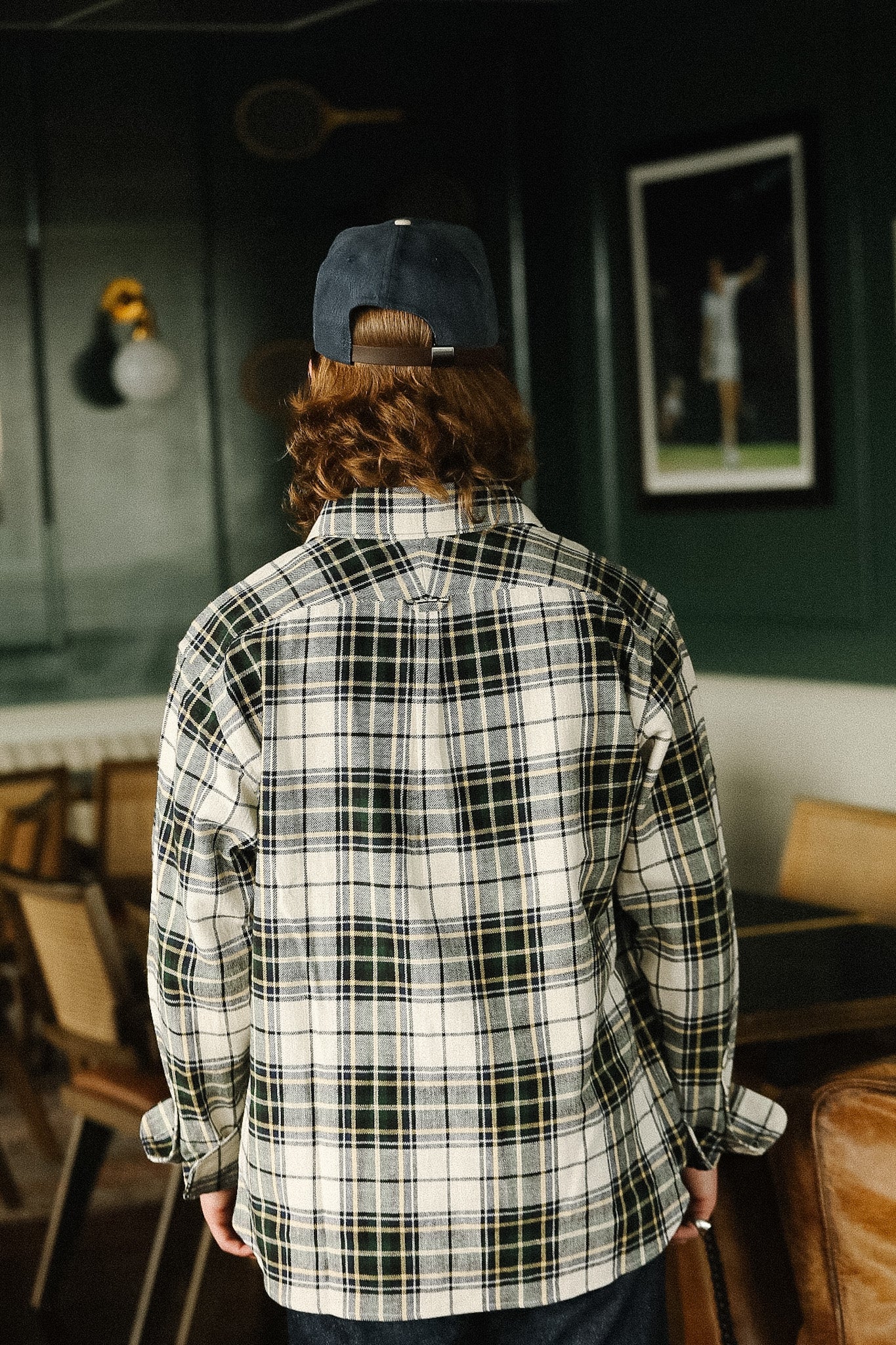 Person wearing a plaid shirt and cap in a room with chairs and a wall-mounted light fixture.