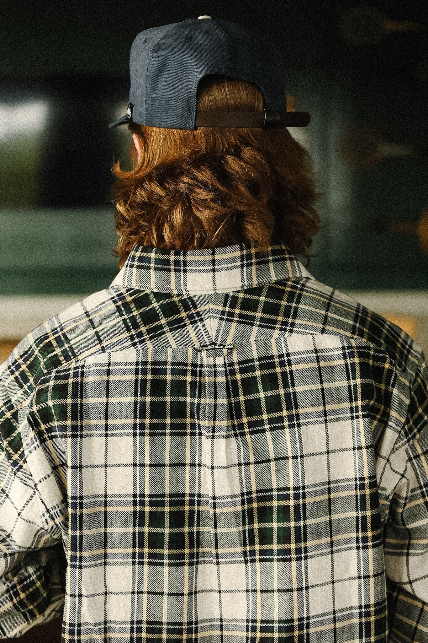 Person wearing a plaid shirt and cap from behind, with a blurred background