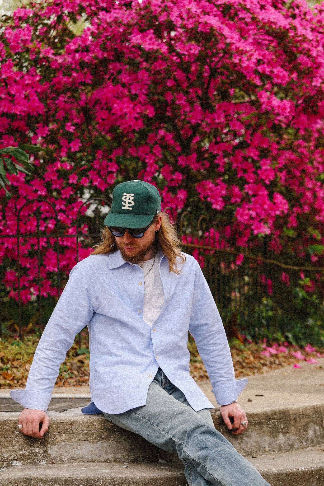 Person wearing a green cap and light blue shirt sitting on steps with pink flowers in the background