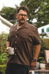 A man wearing a brown short-sleeve button-up shirt and glasses, holding a white mug in an outdoor backyard setting.